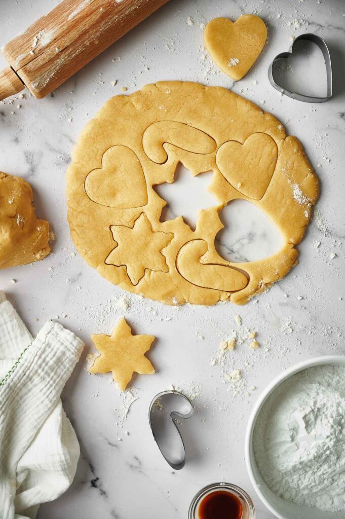 Rolled-out cookie dough on a floured surface with heart, star, and candy cane shapes cut out for Christmas sugar cookies, surrounded by cookie cutters, a rolling pin, a cloth, and a bowl of flour.