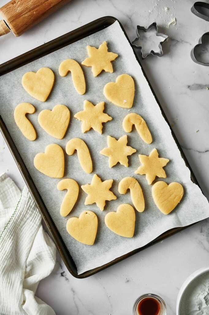 Tray with unbaked Christmas Sugar Cookies in heart, candy cane, and star shapes on parchment paper, surrounded by baking tools on a marble surface.