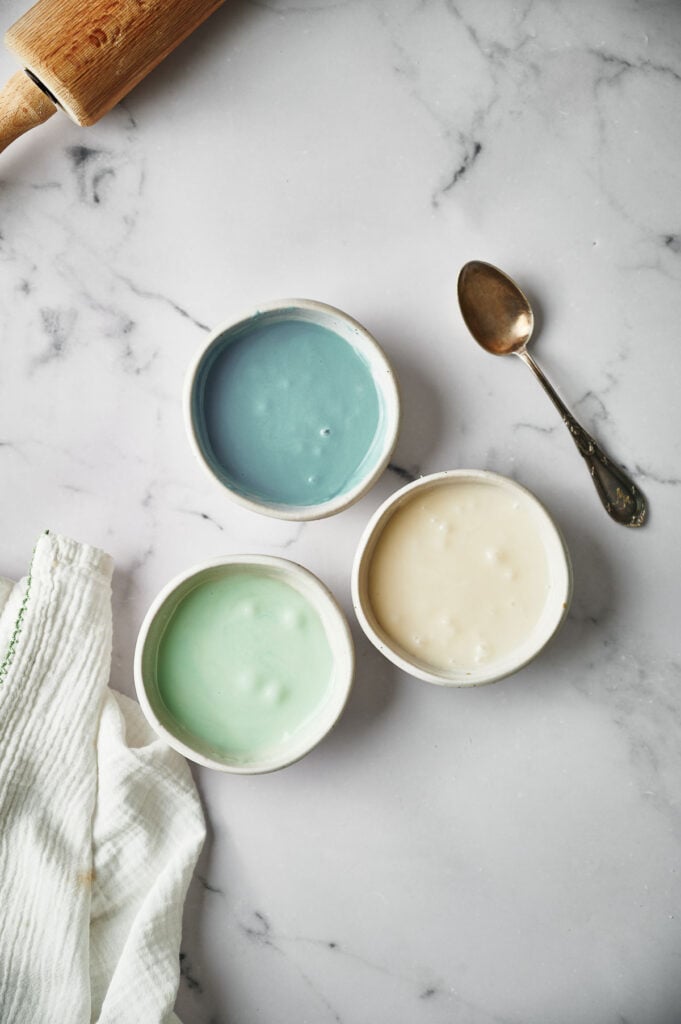 Three small bowls with blue, green, and white icing for decorating Christmas sugar cookies sit on a marble surface, next to a rolling pin, vintage spoon, and a white kitchen towel.
