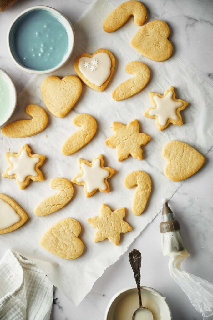 Assorted Christmas sugar cookies in heart, star, and candy cane shapes rest on parchment paper, some decorated with white icing, with bowls of icing and a piping bag nearby.