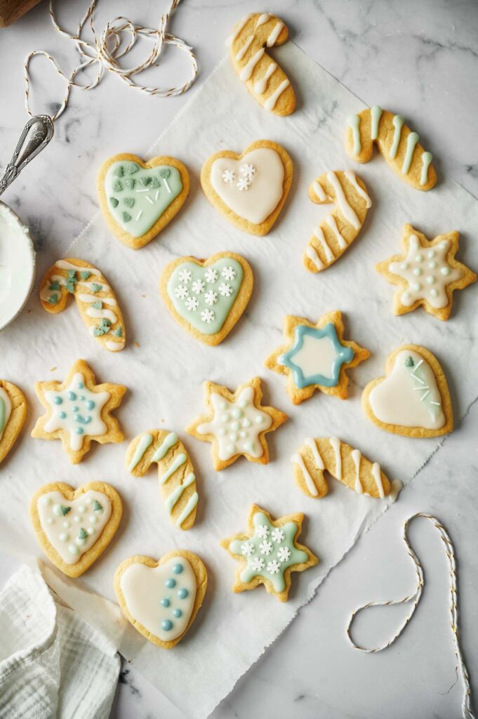 Christmas Sugar Cookies in shapes of hearts, stars, snowflakes, and candy canes are arranged on parchment paper atop a marble surface. String and a cloth sit nearby.