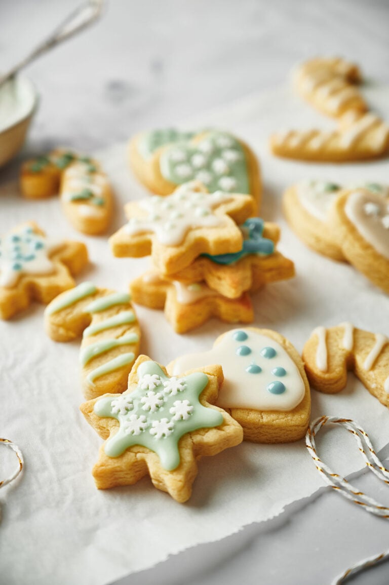 Assorted Christmas Sugar Cookies in various shapes, including snowflakes and mittens, are arranged on a piece of parchment paper on a light surface.