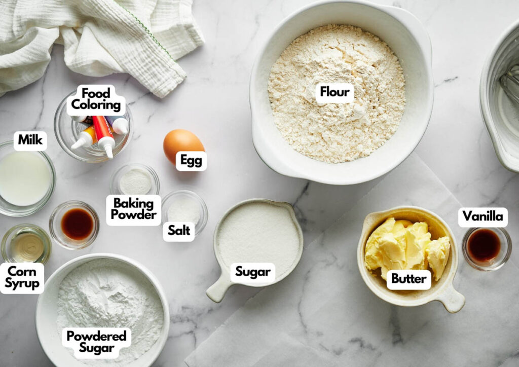 A top-down view of labeled baking ingredients for Christmas Sugar Cookies: flour, sugar, powdered sugar, corn syrup, vanilla, butter, baking powder, salt, food coloring, an egg, and milk on a white surface.
