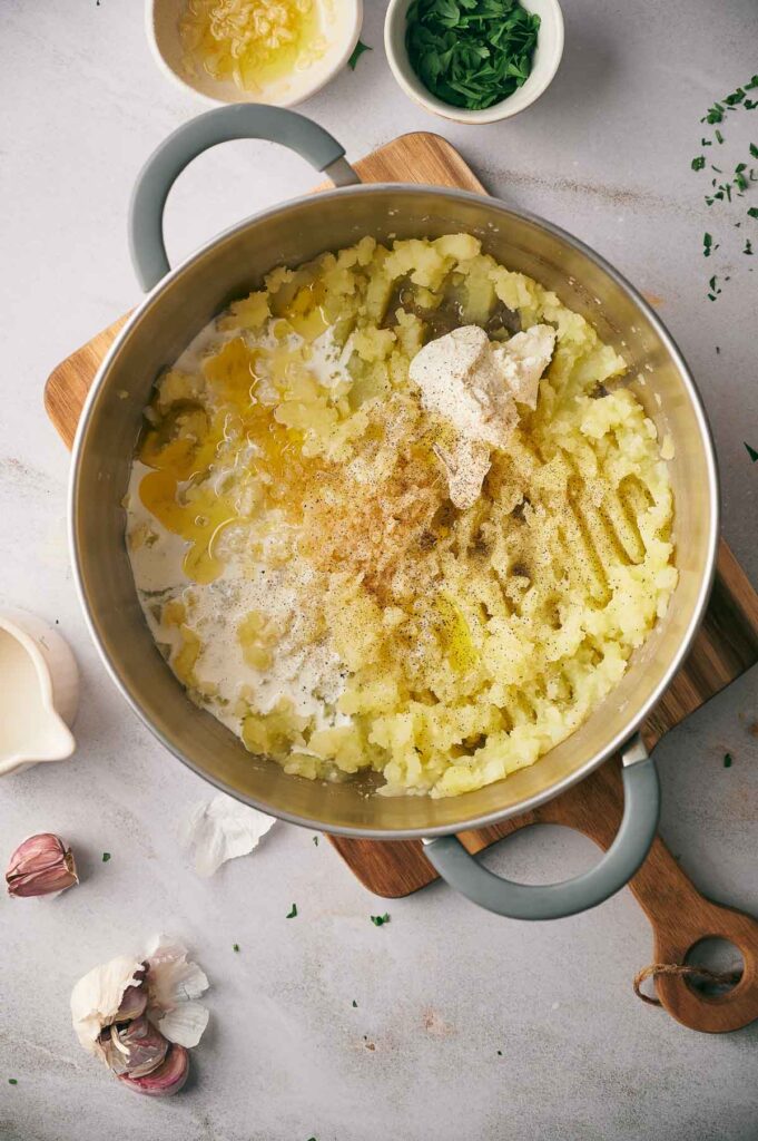 A pot of Southern Mash Potato, made with butter, cream, and spices, sits on a wooden board, surrounded by garlic, parsley, and a small bowl of chopped herbs.