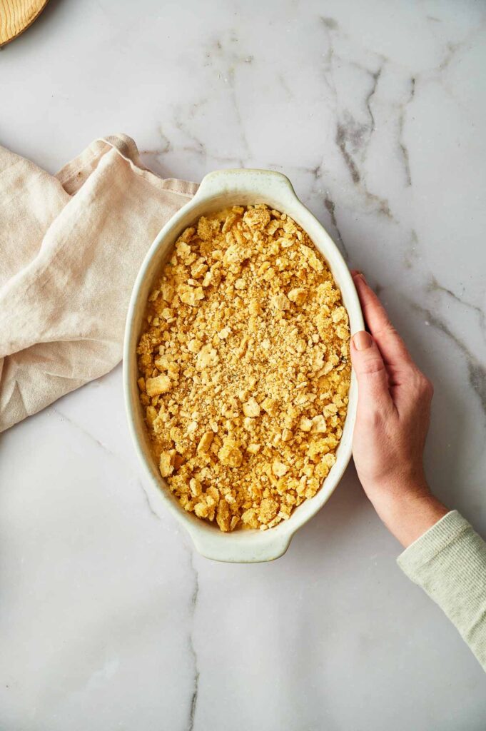 A hand holds an oval baking dish filled with unbaked Yellow Squash Casserole topped with a crumb topping, placed on a marble surface next to a beige cloth.
