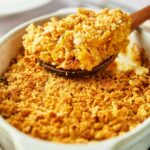 A close-up of a Yellow Squash Casserole with a golden, crumbly topping being served with a wooden spoon from a white dish.