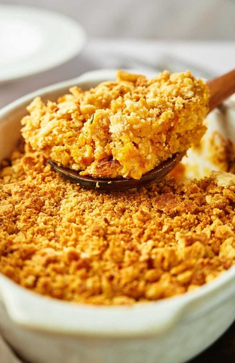 A close-up of a Yellow Squash Casserole with a golden, crumbly topping being served with a wooden spoon from a white dish.