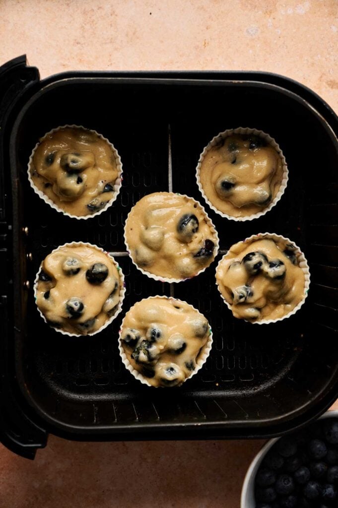 Six silicone muffin cups filled with Air Fryer Blueberry Muffins batter are arranged in an air fryer basket, ready to be baked.