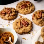 Five Air Fryer Butter Pecan Cookies are arranged on a light surface, with a small bowl of ground cinnamon and a wooden spoon nearby.