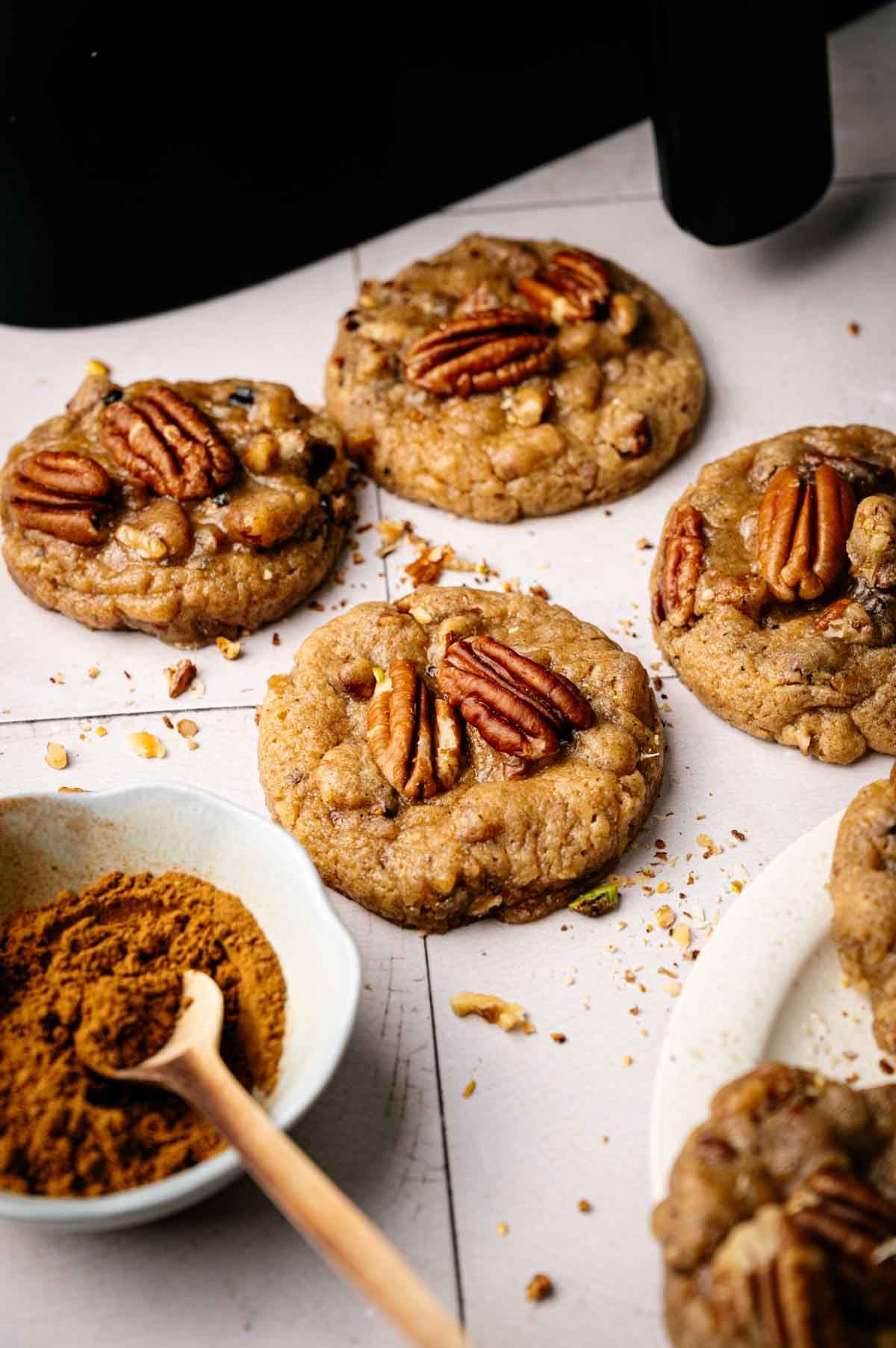 Five Air Fryer Butter Pecan Cookies are arranged on a light surface, with a small bowl of ground cinnamon and a wooden spoon nearby.