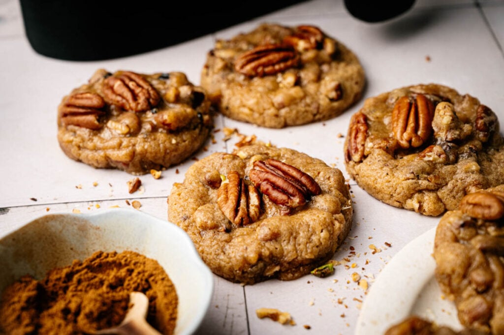 Four Air Fryer Butter Pecan Cookies rest on a white surface, topped with pecans and surrounded by scattered crumbs, with a small bowl of ground spice in the foreground.