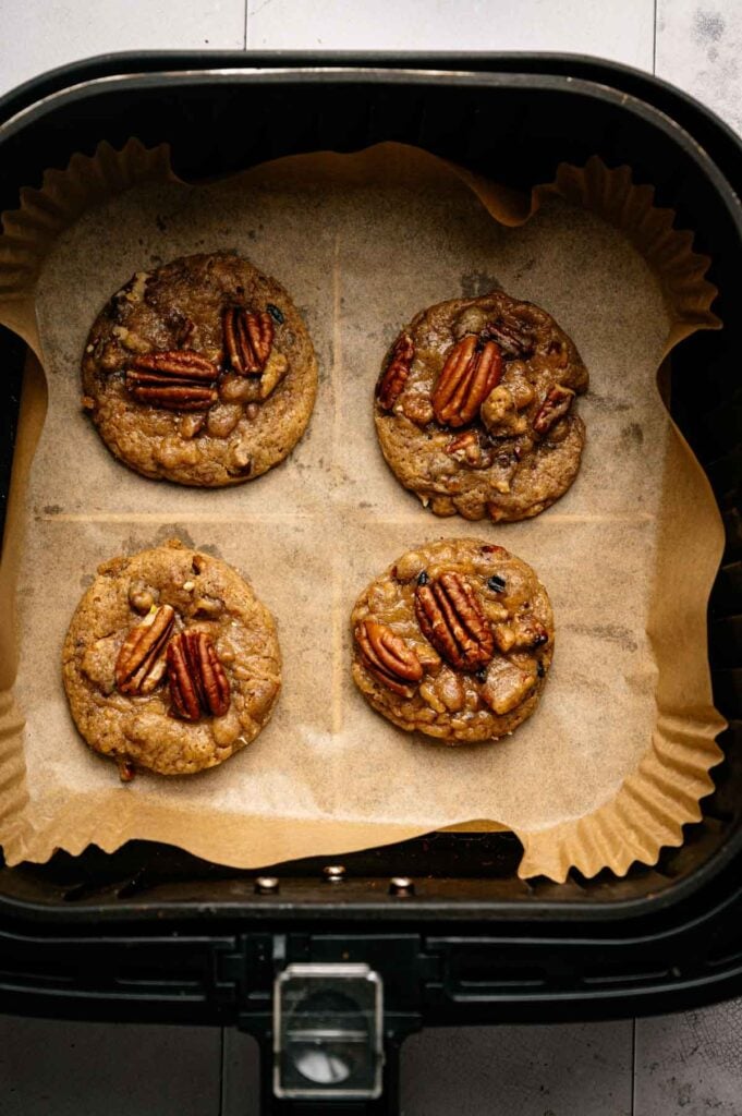 Four Air Fryer Butter Pecan Cookies are baking on parchment paper inside an air fryer basket, each topped with a toasted pecan.