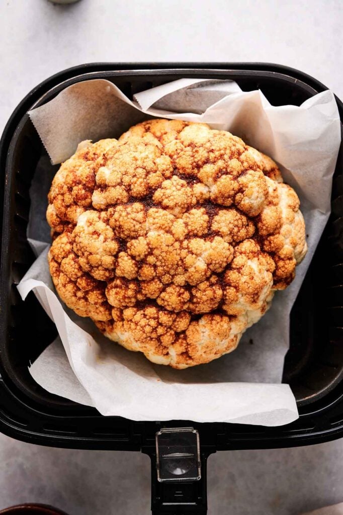 A whole cauliflower head, seasoned with spices, rests on parchment paper in an air fryer basket&mdash;perfect for making Air Fryer Roasted Whole Cauliflower.