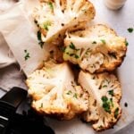 Four Air Fryer Roasted Whole Cauliflower wedges, garnished with chopped parsley, are arranged on parchment paper next to a pepper grinder and an air fryer basket.