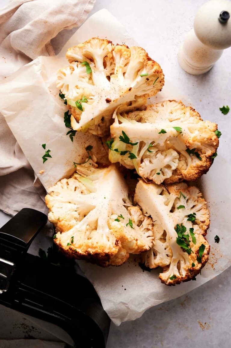Four Air Fryer Roasted Whole Cauliflower wedges, garnished with chopped parsley, are arranged on parchment paper next to a pepper grinder and an air fryer basket.