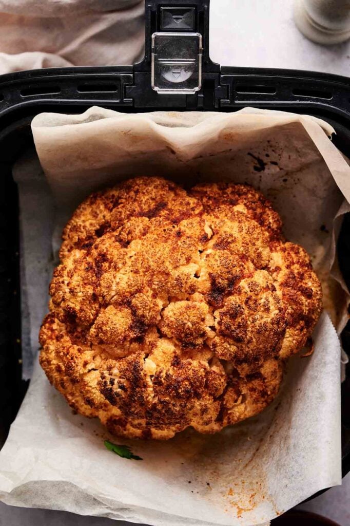 An Air Fryer Roasted Whole Cauliflower with a browned, seasoned crust sits on parchment paper inside an air fryer basket.