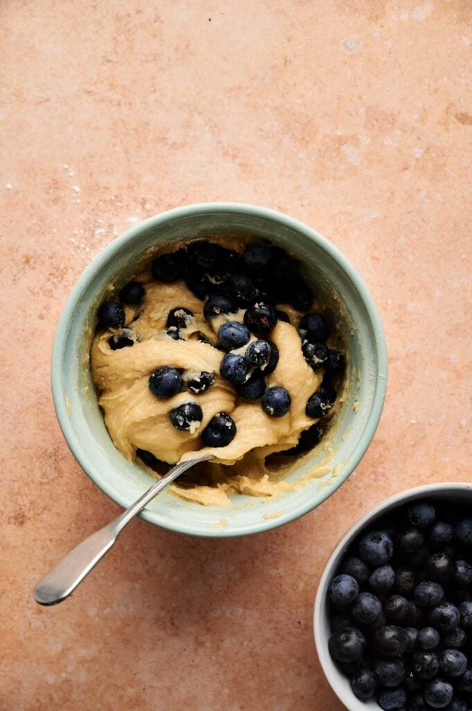 A bowl of muffin batter with fresh blueberries, perfect for Air Fryer Blueberry Muffins, and a spoon beside a smaller bowl filled with blueberries on a light brown surface.