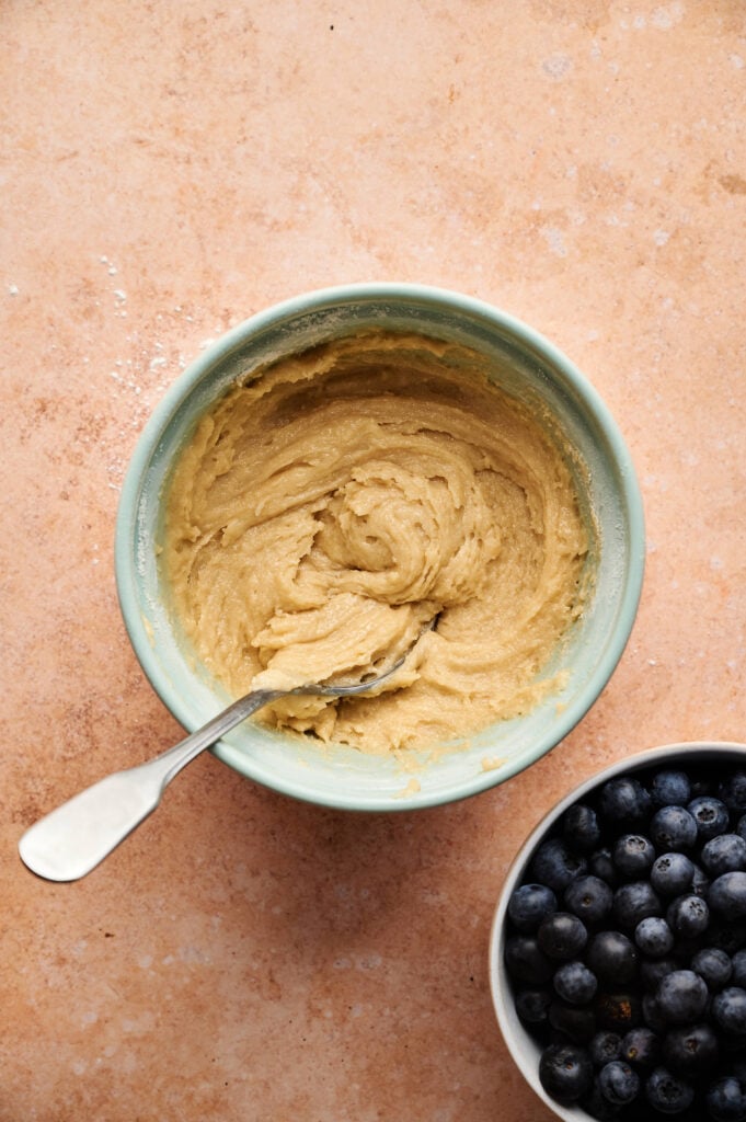 A bowl of light brown Air Fryer Blueberry Muffins batter with a spoon sits next to a bowl of fresh blueberries on a light brown surface.