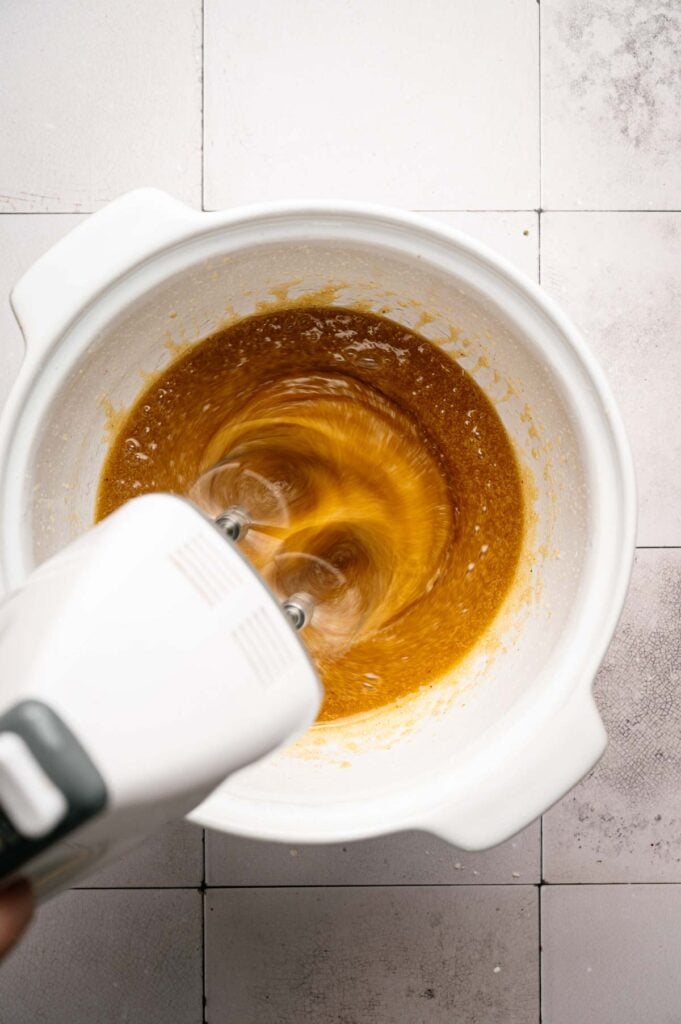 A hand mixer blends a brown liquid mixture in a white mixing bowl on a tiled countertop, preparing dough for Air Fryer Butter Pecan Cookies.