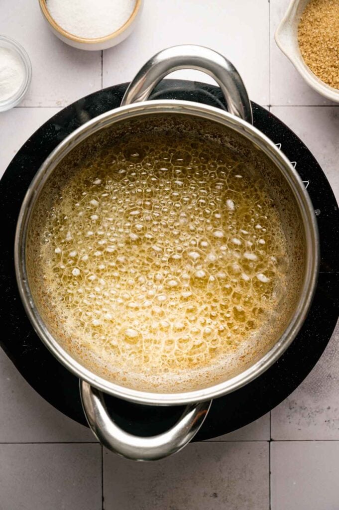 A saucepan of browning butter bubbling on the stovetop, turning deep amber, with bowls of sugar and salt nearby.