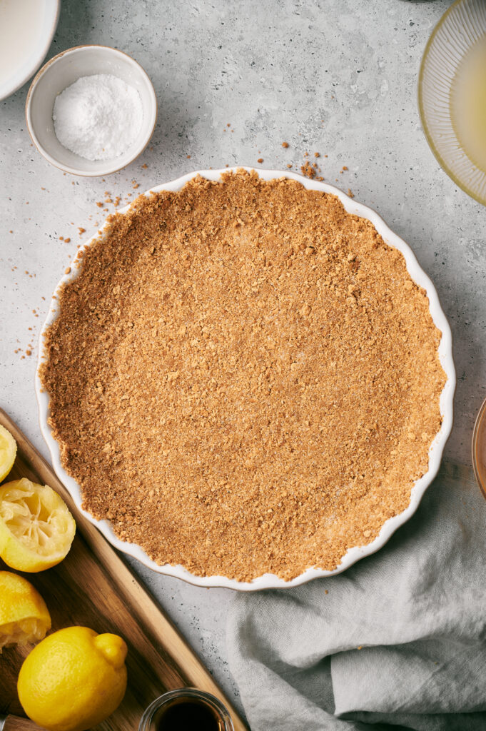 A round dish features a smooth Graham Cracker Crust, surrounded by a lemon, a small bowl of sugar, and a bowl of liquid, all arranged on a light surface.