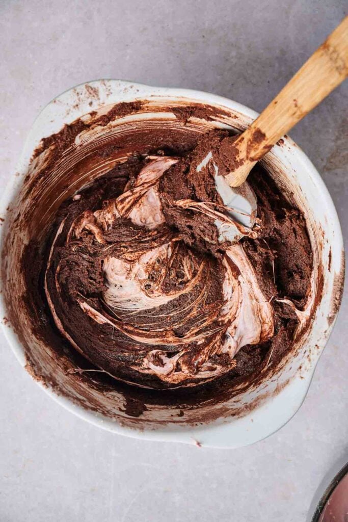 A mixing bowl with chocolate batter and a spatula, showing swirls of white and brown ingredients&mdash;perfect for making Air Fryer Marshmallow Swirl Cookies&mdash;on a light countertop.
