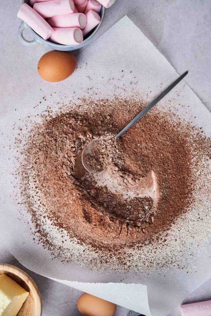 Cocoa powder and flour mixture on parchment paper with a spoon, surrounded by eggs, butter, and a bowl of pink marshmallows&mdash;perfect ingredients for Air Fryer Marshmallow Swirl Cookies.