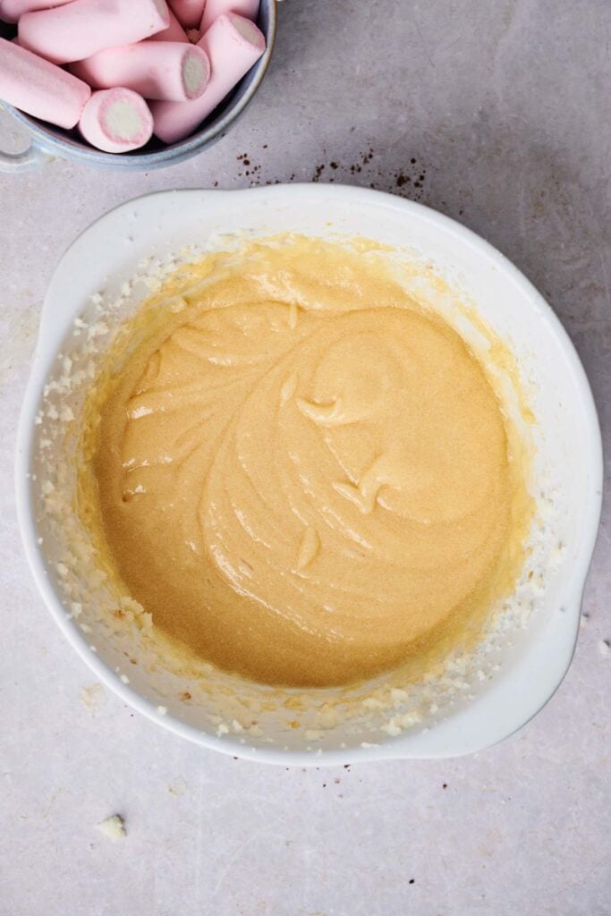 A white mixing bowl containing a smooth, light yellow batter sits on a countertop next to a bowl of pink marshmallows, perfect for making Air Fryer Marshmallow Swirl Cookies.