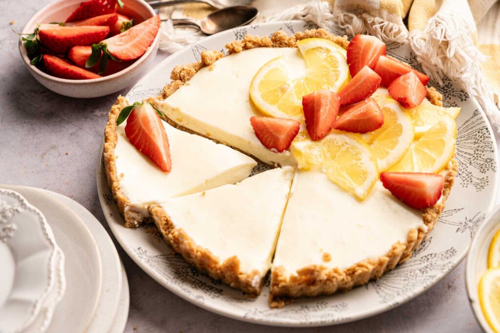 A sliced No Bake Lemon Tart topped with fresh strawberries and lemon slices sits on a plate, with a bowl of strawberries and dessert plates nearby.