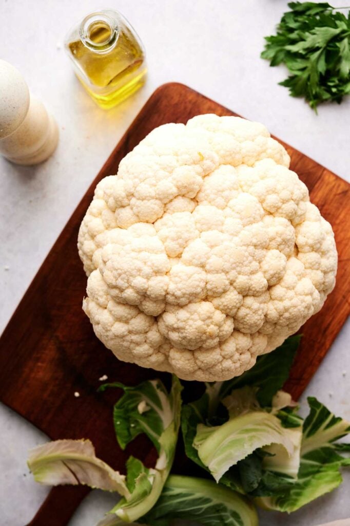 An Air Fryer Roasted Whole Cauliflower sits on a wooden cutting board with some leaves, next to a bottle of olive oil, fresh parsley, and a pepper grinder.