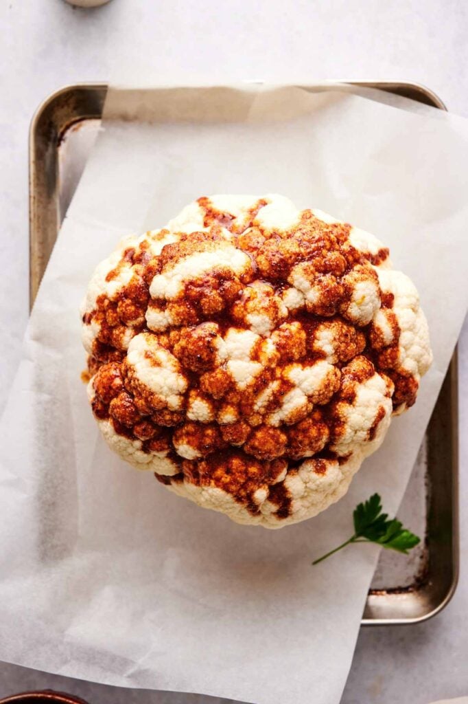 An Air Fryer Roasted Whole Cauliflower sits on a parchment-lined tray, coated with a vibrant reddish-brown spice paste, viewed from above.