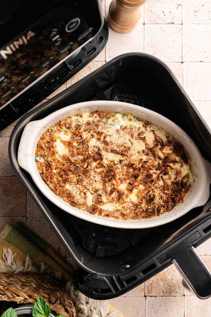 Oval casserole dish filled with Air Fryer Spinach Casserole, topped with a golden browned layer, nestled in an air fryer basket on a tiled surface.