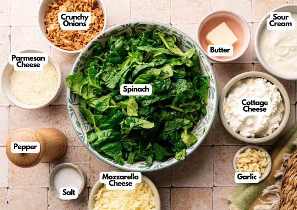 A bowl of chopped spinach surrounded by labeled bowls of crunchy onions, butter, sour cream, cottage cheese, garlic, mozzarella cheese, parmesan cheese, salt, and pepper—all you need for a delicious Air Fryer Spinach Casserole.
