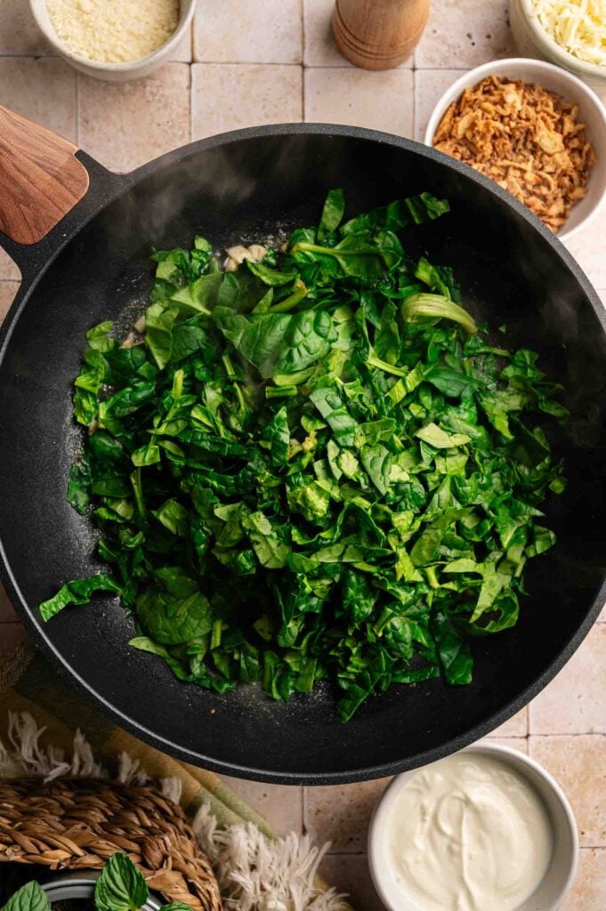 Chopped spinach cooking in a black skillet on a tiled surface, surrounded by bowls of grated cheese, crispy onions, and creamy sauce—ideal ingredients for an Air Fryer Spinach Casserole.