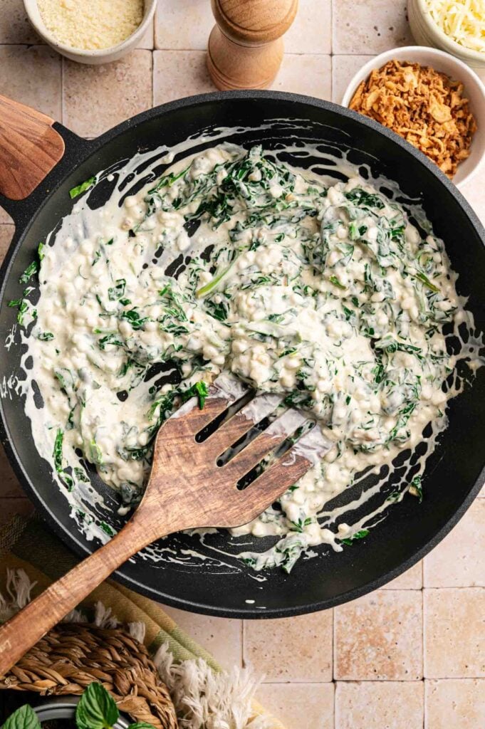 A wooden spoon stirs creamy Air Fryer Spinach Casserole in a black skillet, with bowls of shredded cheese and crispy onions nearby.