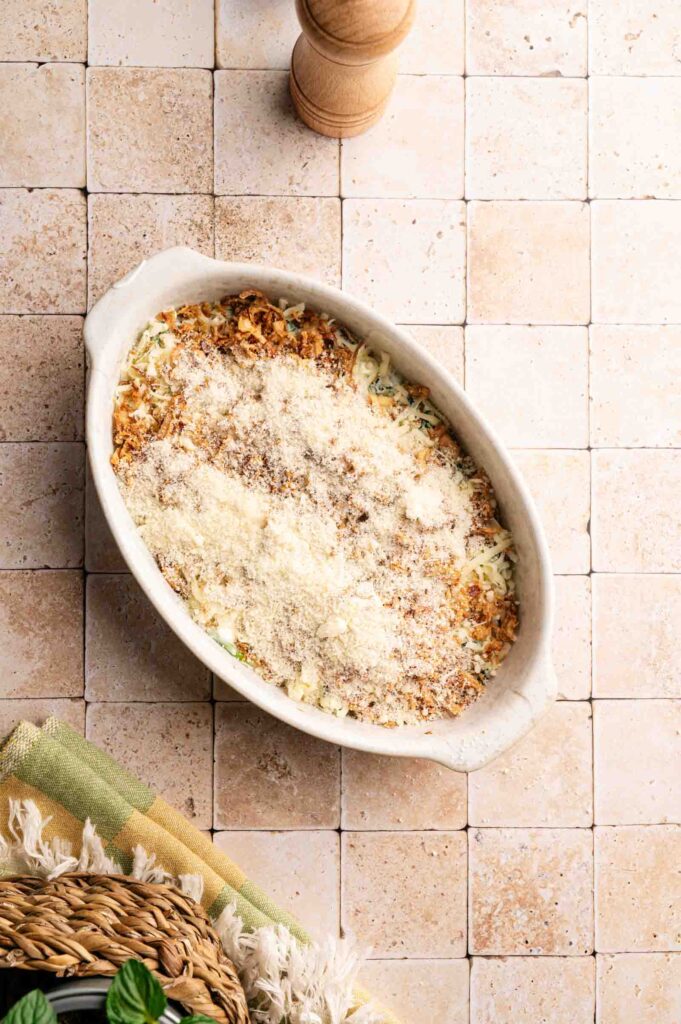 A white oval baking dish filled with spinach casserole mixture, topped with shredded mozzarella, Parmesan, and crispy onions, ready for the air fryer.