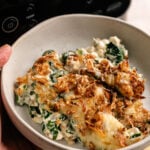 A hand holds a bowl of creamy Air Fryer Spinach Casserole topped with crispy fried onions, in front of a Ninja kitchen appliance.