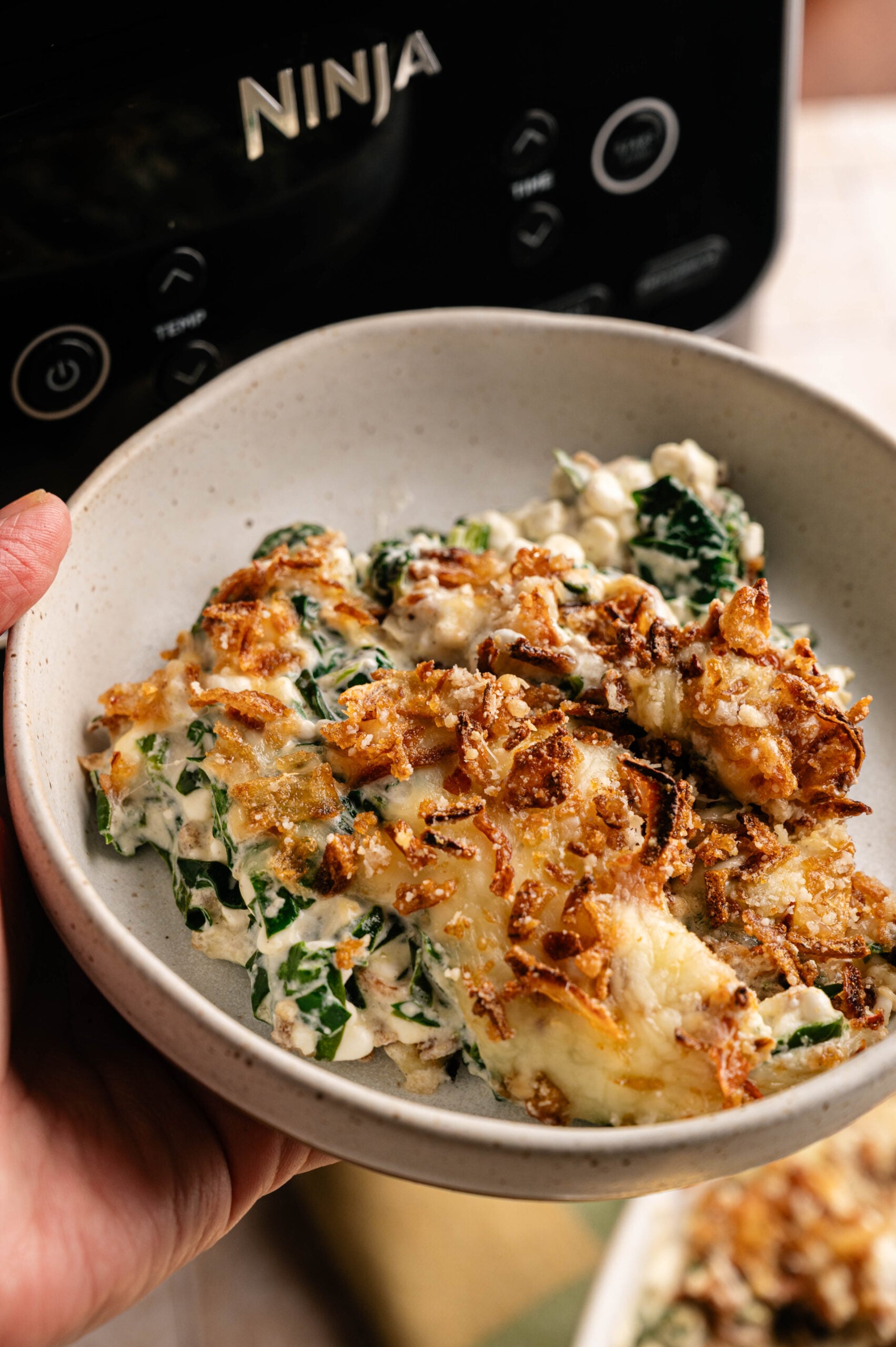 A hand holds a bowl of creamy Air Fryer Spinach Casserole topped with crispy fried onions, in front of a Ninja kitchen appliance.
