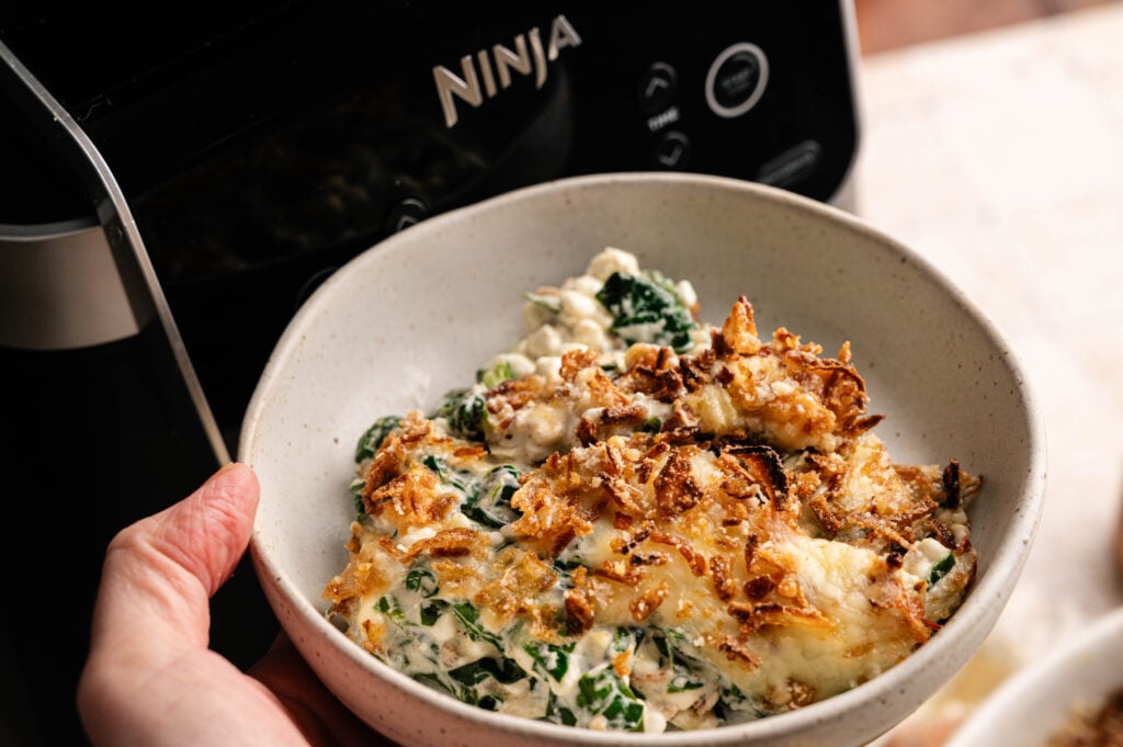 A hand holds a bowl of Air Fryer Spinach Casserole topped with crispy onions, with a Ninja air fryer visible in the background.