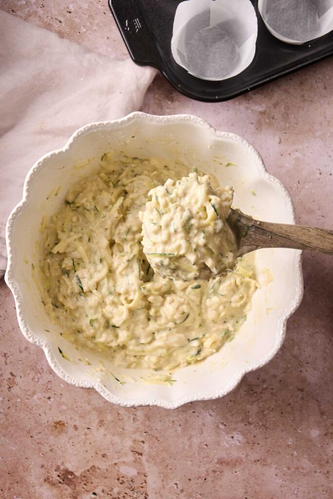 A white bowl filled with thick, mixed batter and shredded vegetables for Air Fryer Zucchini Muffins, with a wooden spoon inside. A muffin tray lined with parchment paper is partially visible nearby.