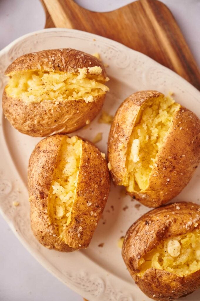 Four baked potatoes with salted, crispy skins are split open and served on a white oval plate, revealing the classic fluffy baked potato interiors melting with butter.