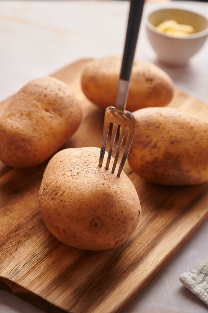 A fork pierces a raw potato on a wooden cutting board with three other potatoes nearby, hinting at the start of a delicious baked potato recipe; a bowl of butter sits in the background.