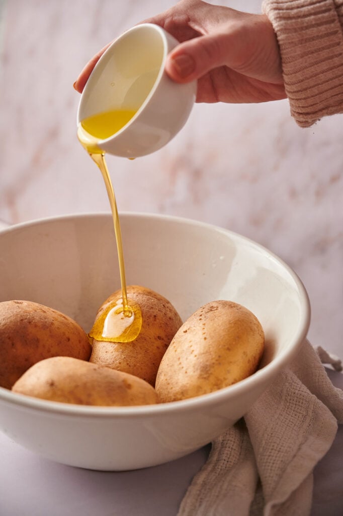 A hand pours oil from a small white cup onto several unpeeled potatoes in a large white bowl, preparing them for baked potato perfection.