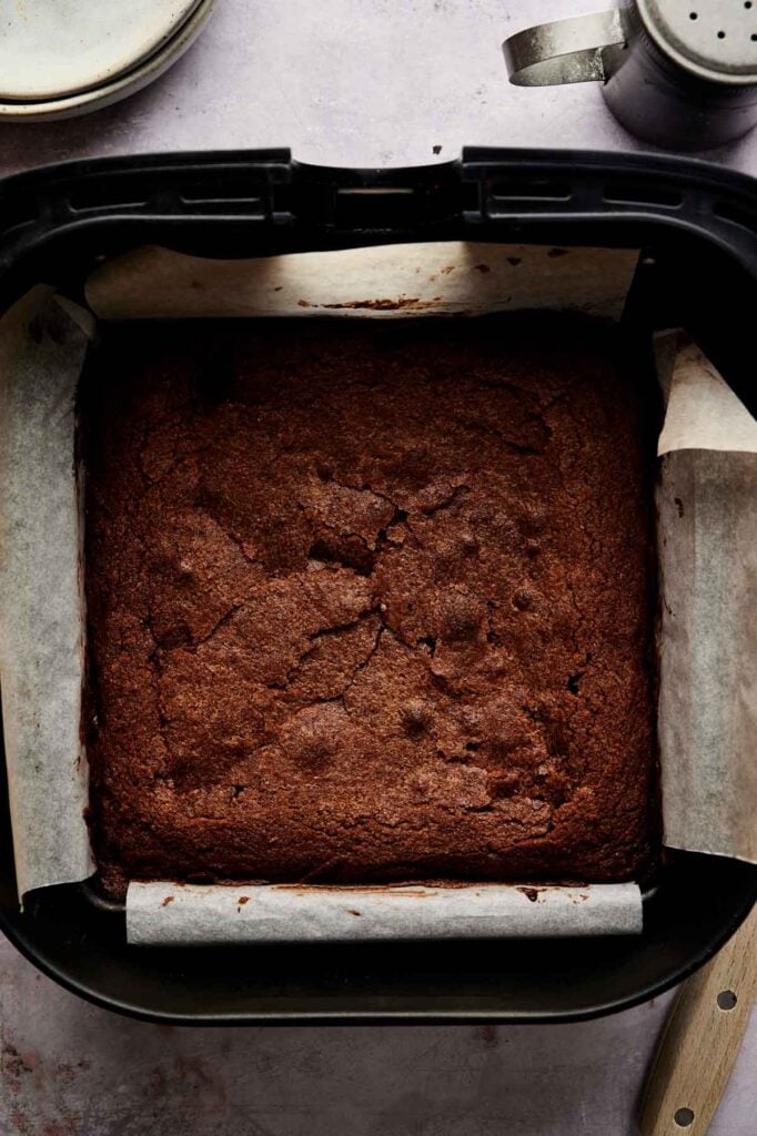 A square Air Fryer Cream Cheese Brownie in a parchment-lined baking pan, shown fresh out of the oven on a light countertop.
