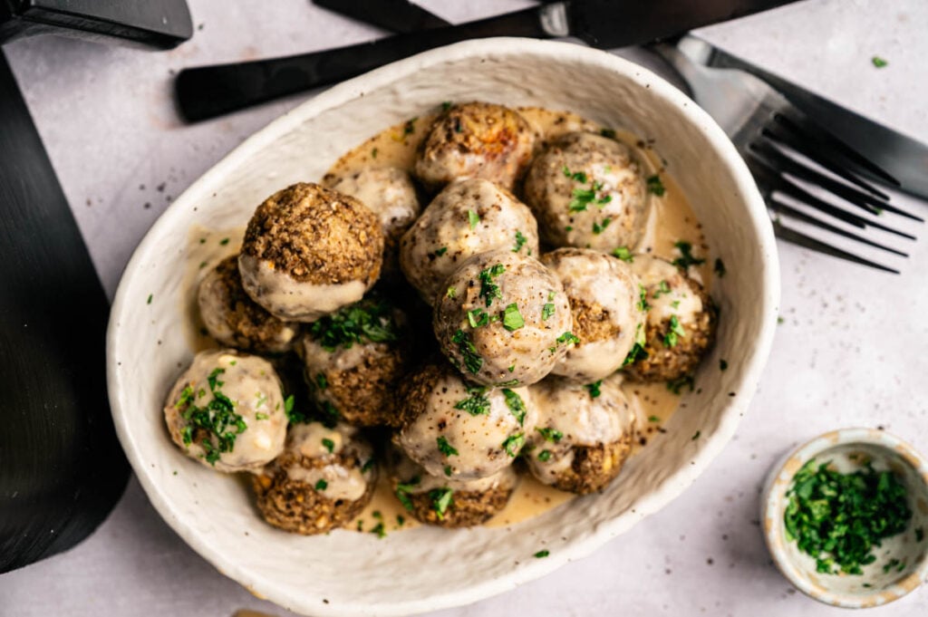 A bowl of Air Fryer Vegetarian Meatballs covered in creamy sauce, garnished with chopped parsley, with utensils and a small dish of herbs nearby.