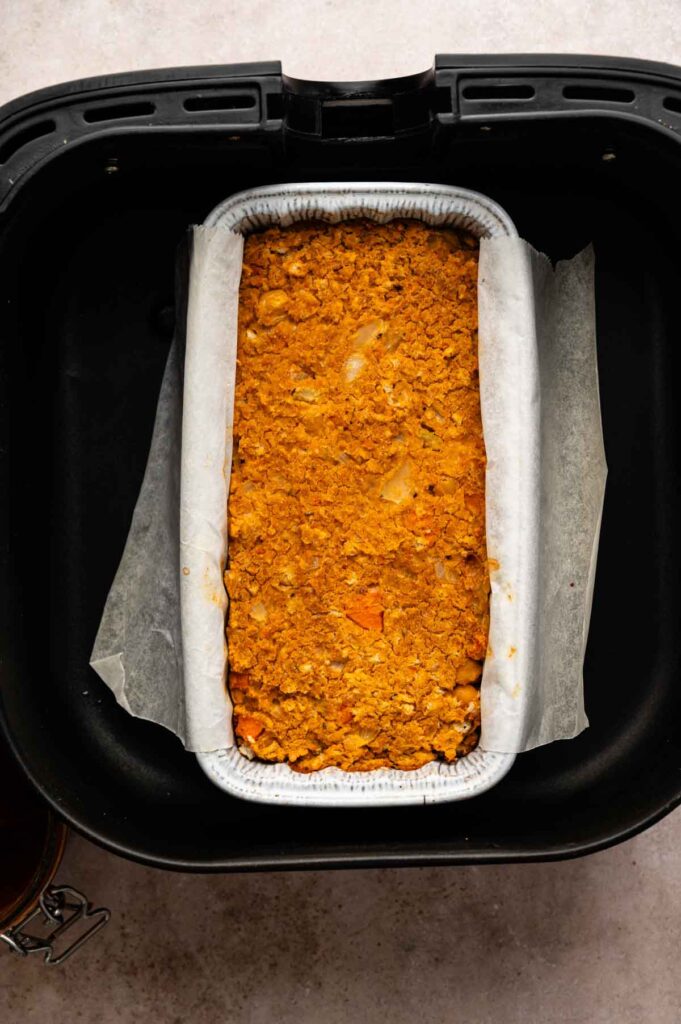 A rectangular loaf pan lined with parchment paper holds an orange-brown vegetarian meatloaf mixture, placed inside an air fryer basket.