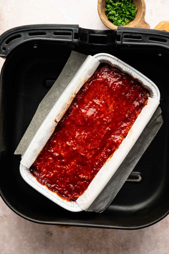 An Air Fryer Vegetarian Meatloaf sits glazed in a parchment-lined loaf pan inside the air fryer basket, with chopped herbs in a small wooden bowl nearby.