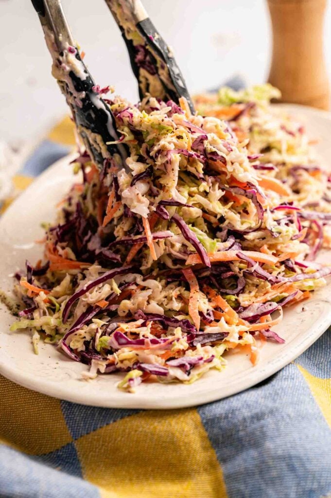Tongs lifting a serving of vibrant coleslaw, filled with shredded cabbage and carrots, onto a white plate, set atop a blue and yellow cloth.