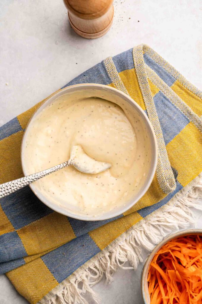 A bowl of creamy coleslaw dressing with a spoon rests on a yellow and blue checkered cloth, with shredded carrots and a pepper grinder nearby.