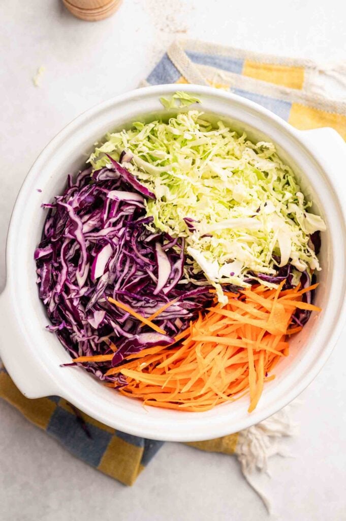 A white bowl contains shredded green cabbage, purple cabbage, and carrots&mdash;classic coleslaw ingredients&mdash;arranged in separate sections on a kitchen towel.